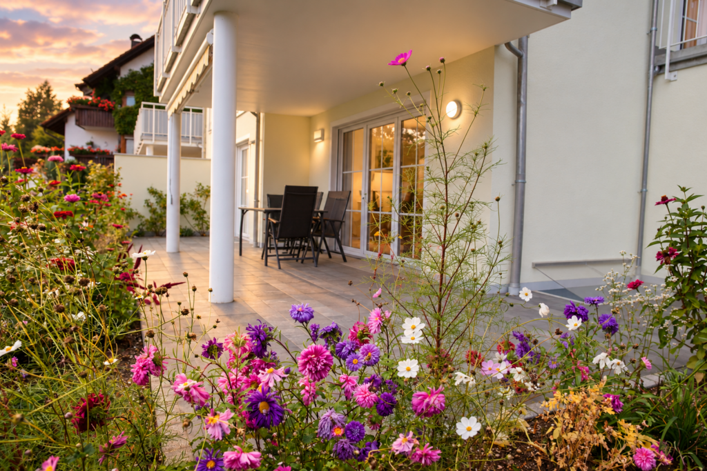 Blühende Stauden vor einer Terrasse mit dunklen Gartenstühlen, weißen Stützen und bodentiefen Türen